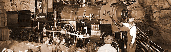 A man stands in front of a large antique railroad engine.